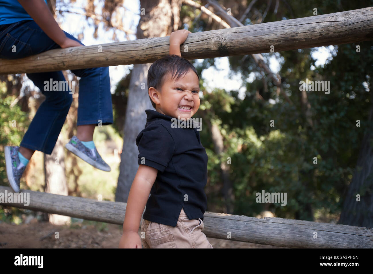 A happy little brother who wants to climb up a fence to sit together with his older sister in a woodland park. Stock Photo