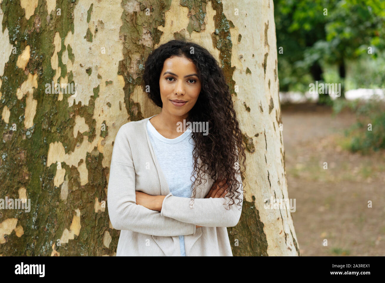 Thoughtful young woman with a quiet smile standing with folded arms against the trunk of a large tree outdoors in a park Stock Photo