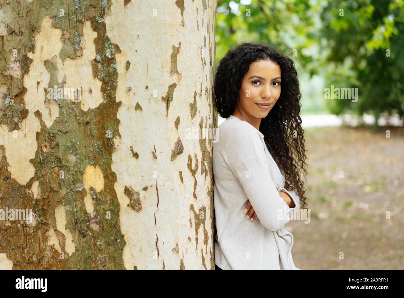Smiling young woman outdoors in a park standing leaning against a large tree trunk with folded arms looking at the camera Stock Photo