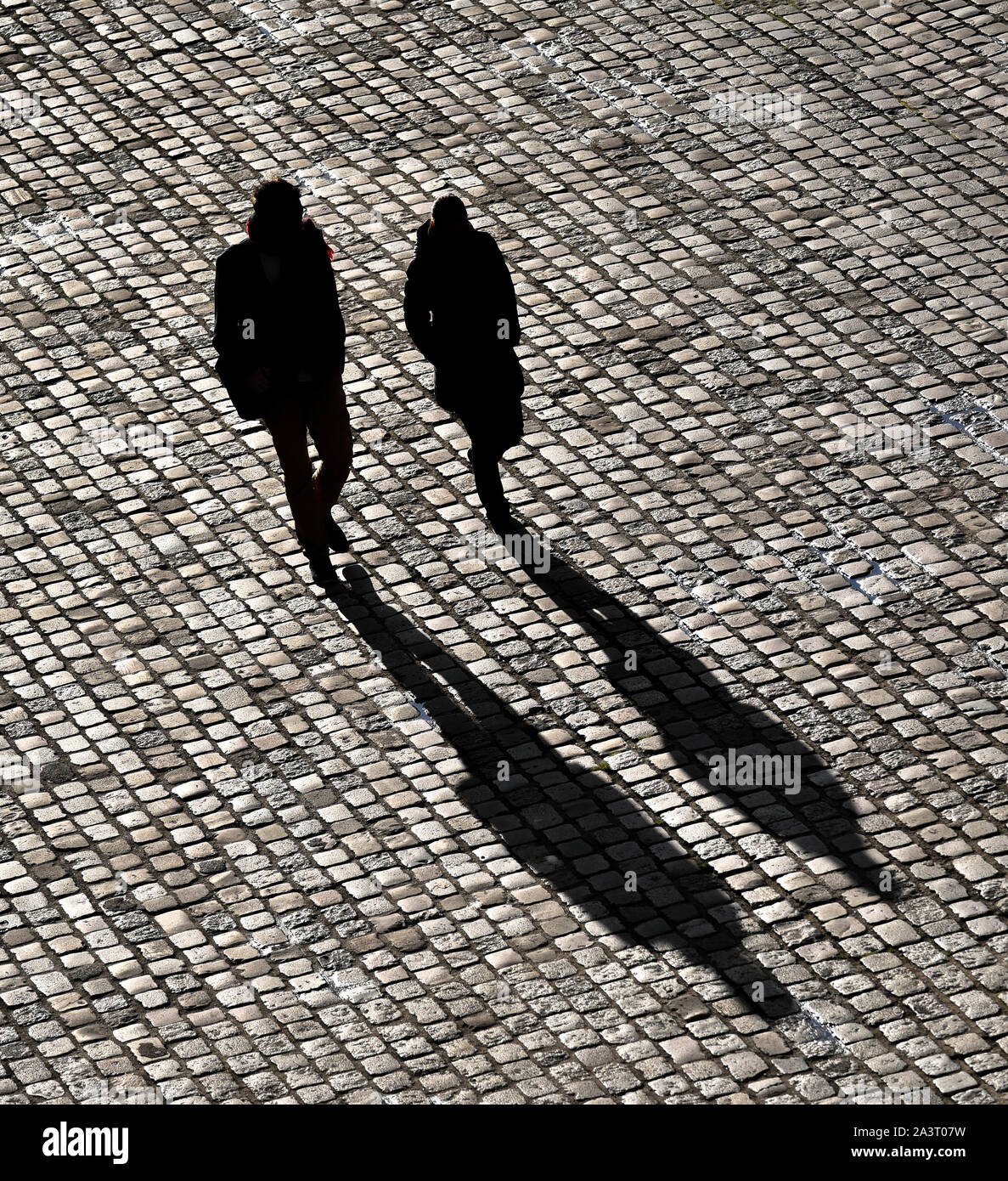 Two bystanders walking on a paved road, projecting their shadows. Stock Photo