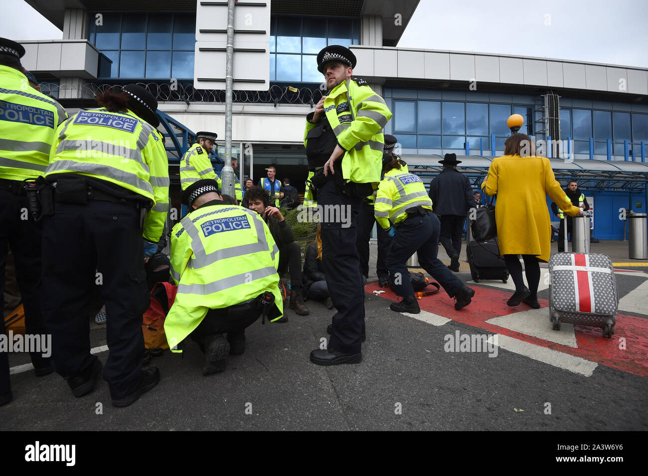 Passengers walk past with suitcases as police search protesters, after activists staged a 'Hong Kong style' blockage of the exit from the train station to City Airport, London, during an Extinction Rebellion climate change protest. Stock Photo