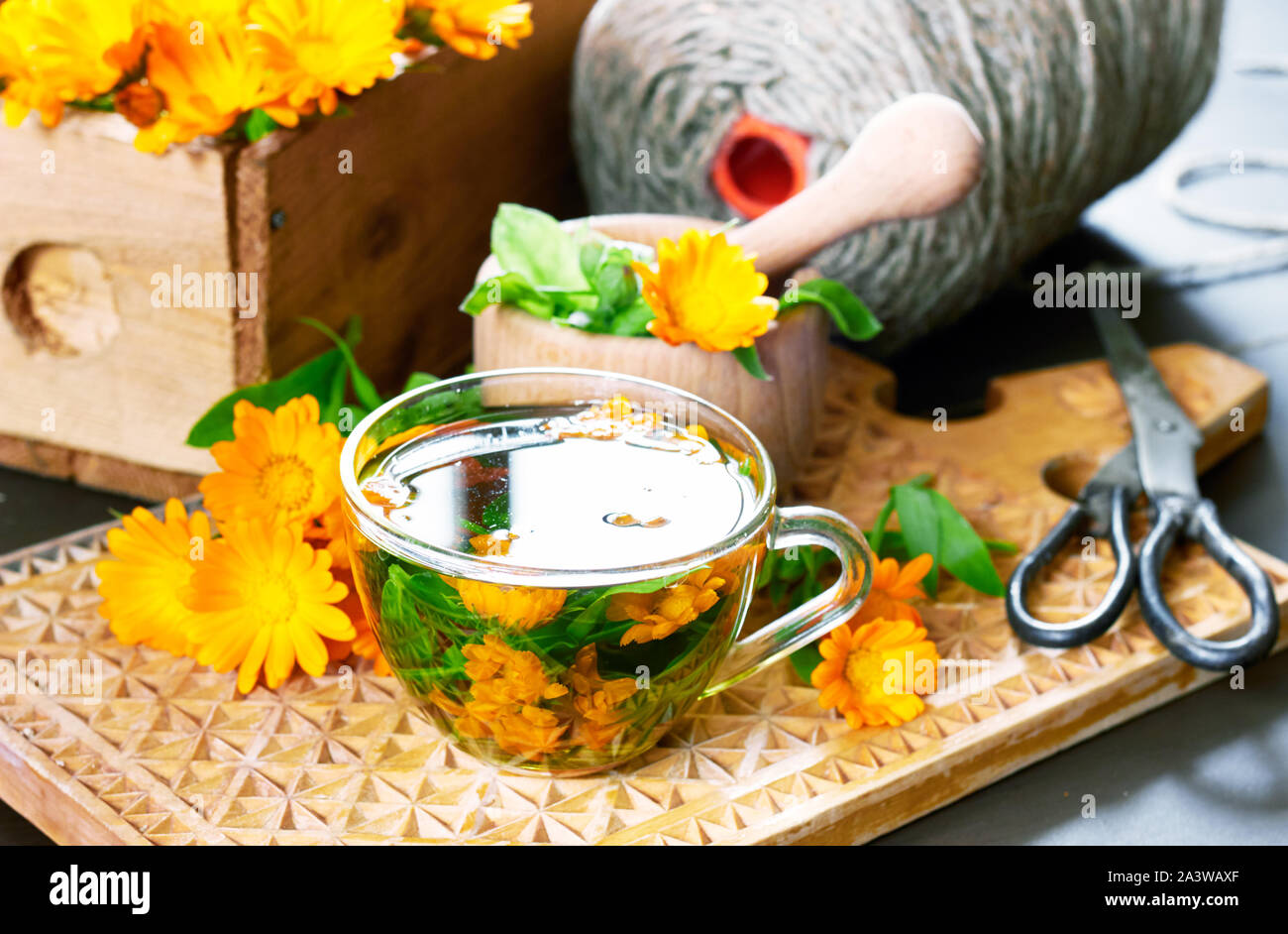 A cup of calendula tea, herbal healing drink made of bunch of fresh flowers with marigold bouquet nearby, a mortar with pounder,  wooden box with cut Stock Photo