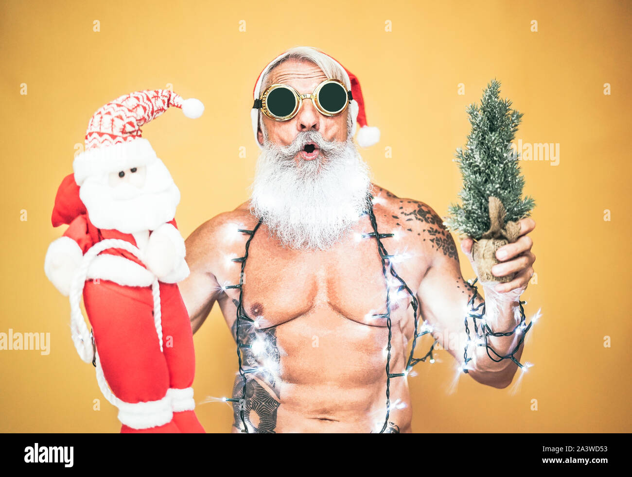 Happy fit santa claus equiped with white christmas lights - Trendy beard hipster senior holding a mini santa claus puppet and xmas tree Stock Photo