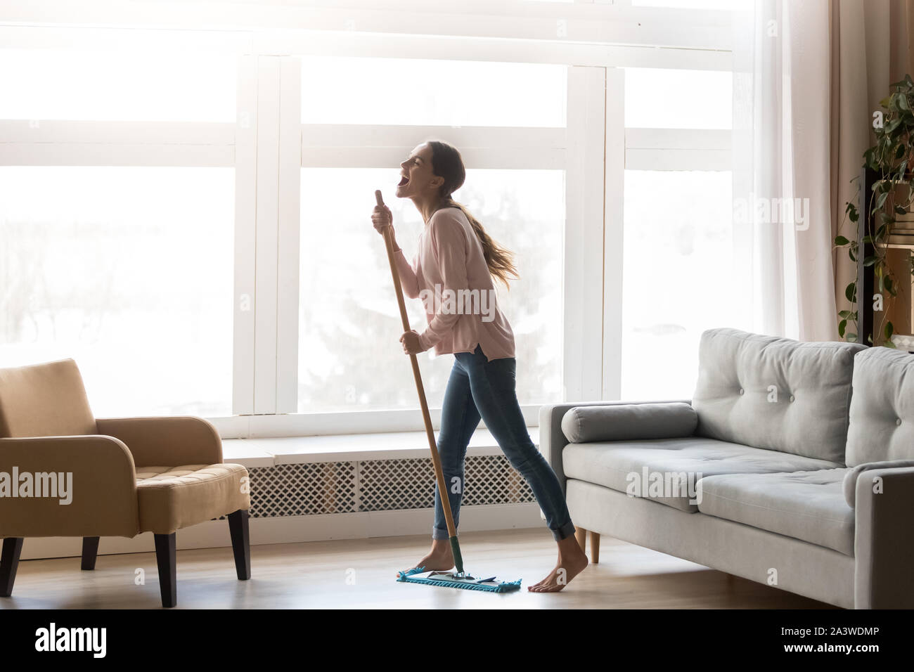 Funny woman holding mop microphone singing cleaning in living room Stock Photo