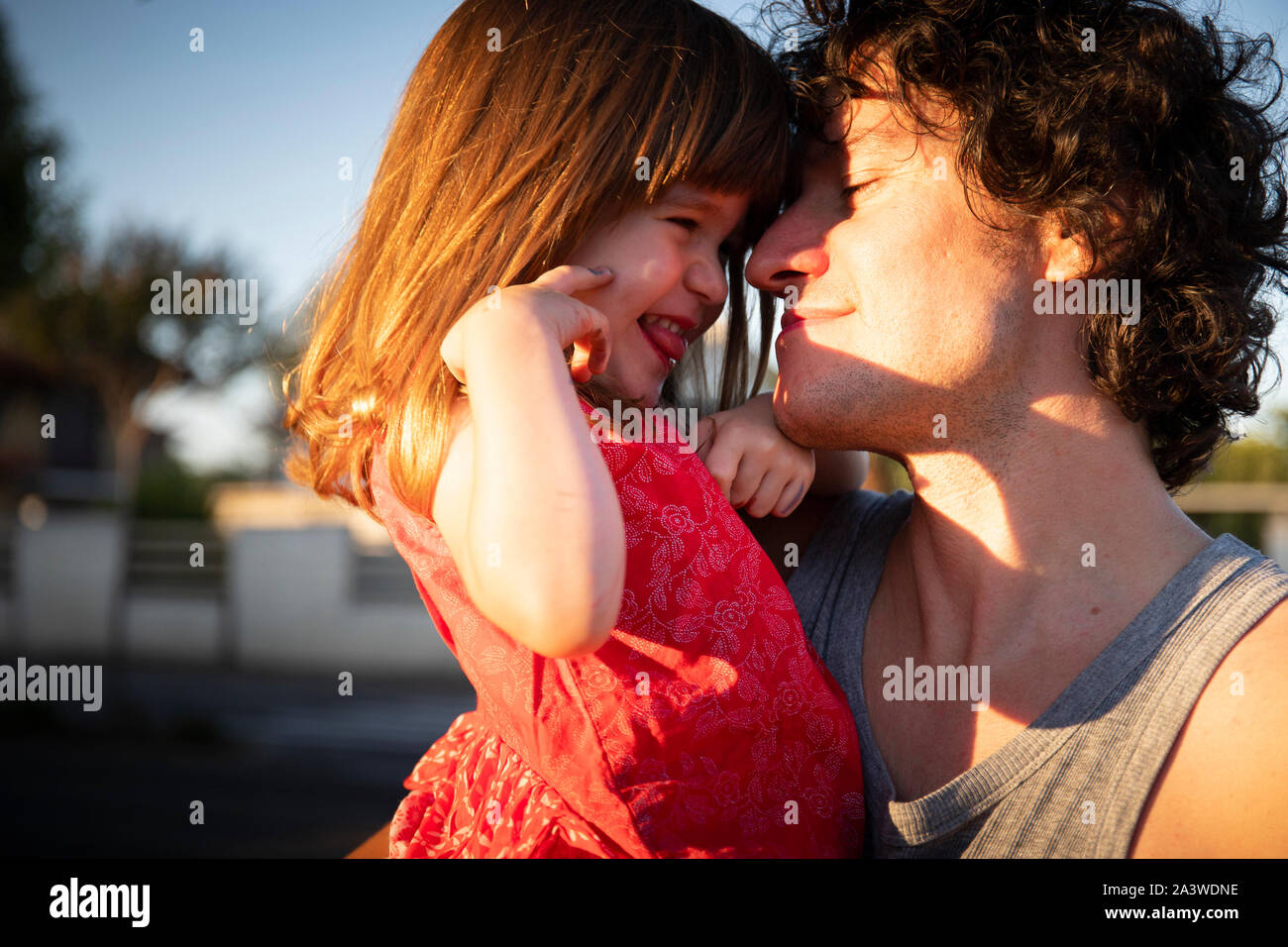 Young father laughing with his little daughter outdoor at sunset. The man is looking at the child and she is wearing a red dress. Copy space on the le Stock Photo