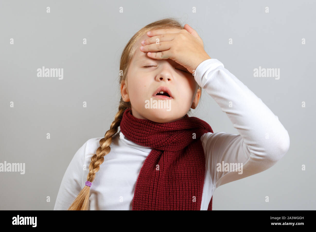 A little girl in a scarf is suffering from an illness. The child holds on to his head. The concept of the season of colds. Stock Photo