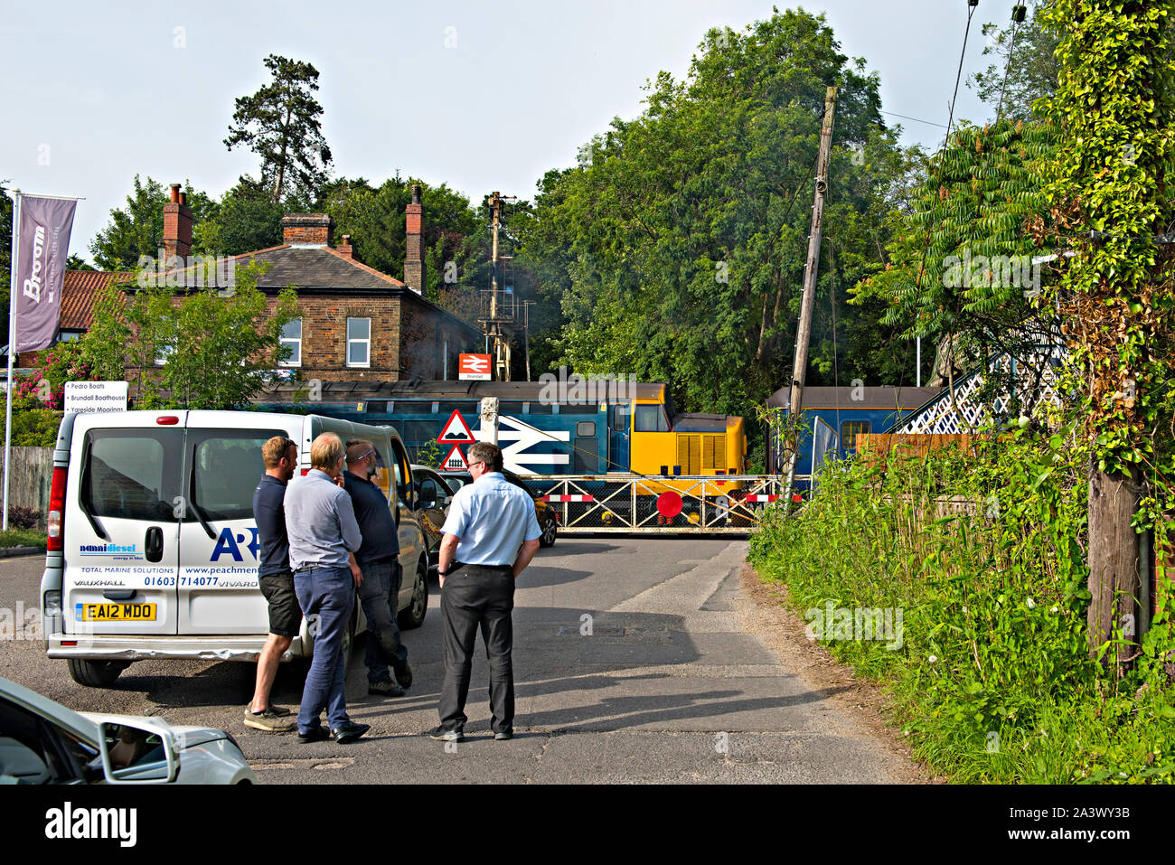Traffic waits at the level crossing at Brundall Railway Station, Norfolk, UK. A class 37 diesel locomotive passes the crossing. Stock Photo