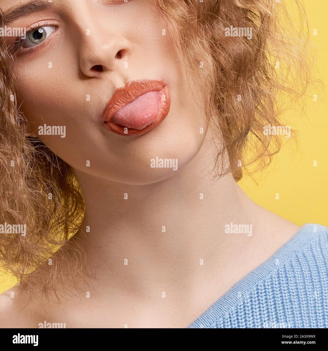 Closeup of bright young woman looking at camera and showing tongue on yellow isolated background. Short haired curly girl posing and smiling in studio. Concept of happiness and children. Stock Photo