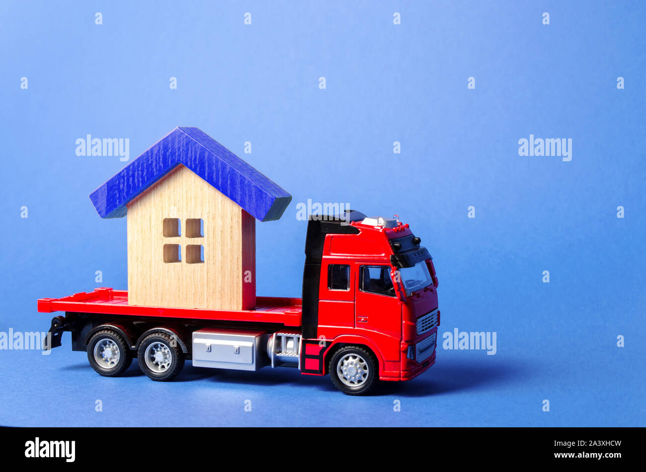 Red truck carrier transports a blue roofed house. Concept of transportation and cargo shipping, moving company. Construction of new houses and objects Stock Photo