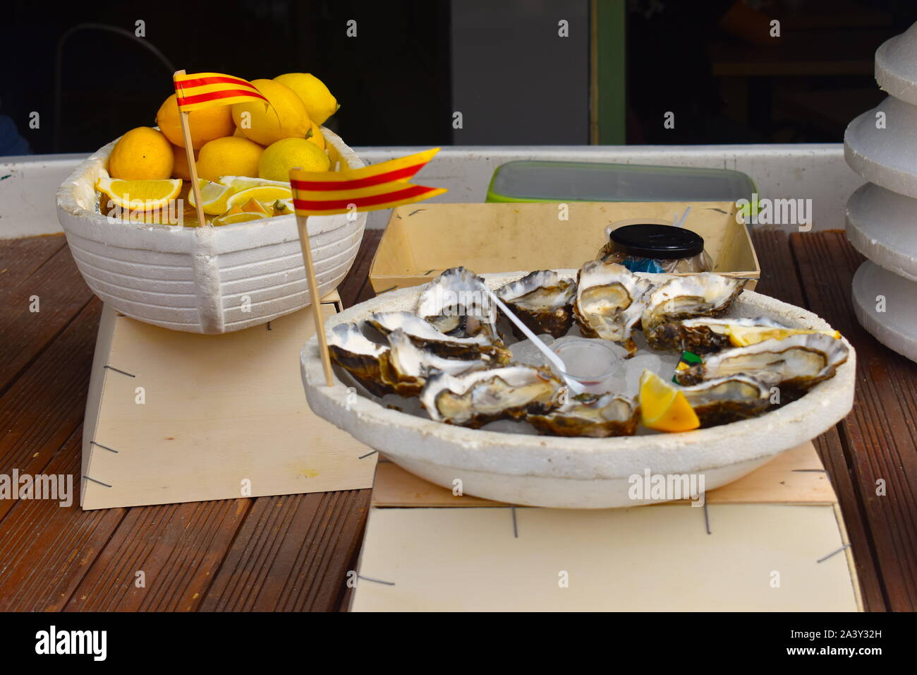 Plate full of French oysters for sale on the local market in the town centre. Bowl full of slices of lemon and Catalan flags standing out. Stock Photo