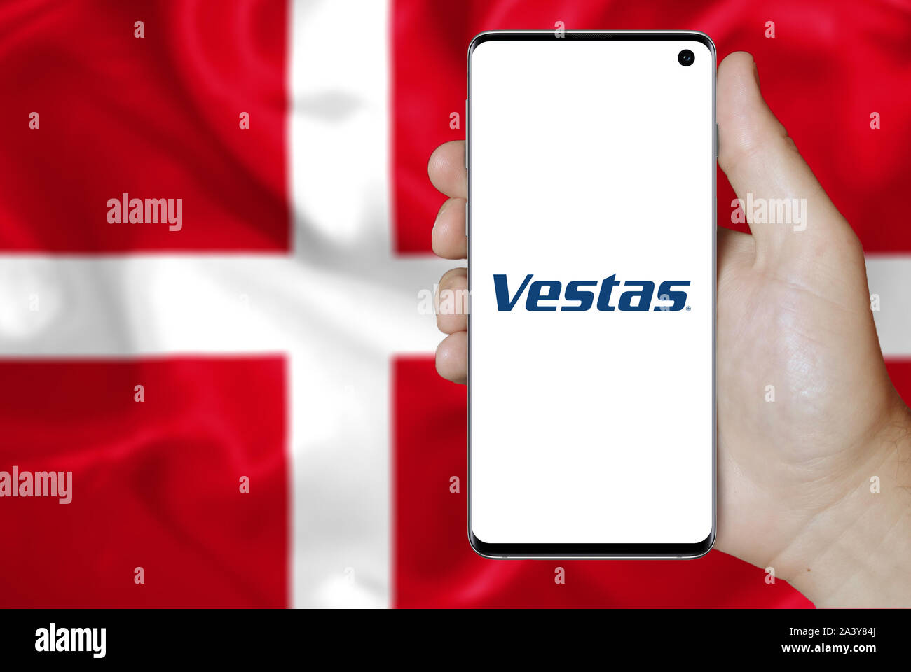 A man holds a phone displaying the logo of company Vestas Wind Systems listed on OMX Copenhagen. Danish flag background. Credit: PIXDUCE Stock Photo