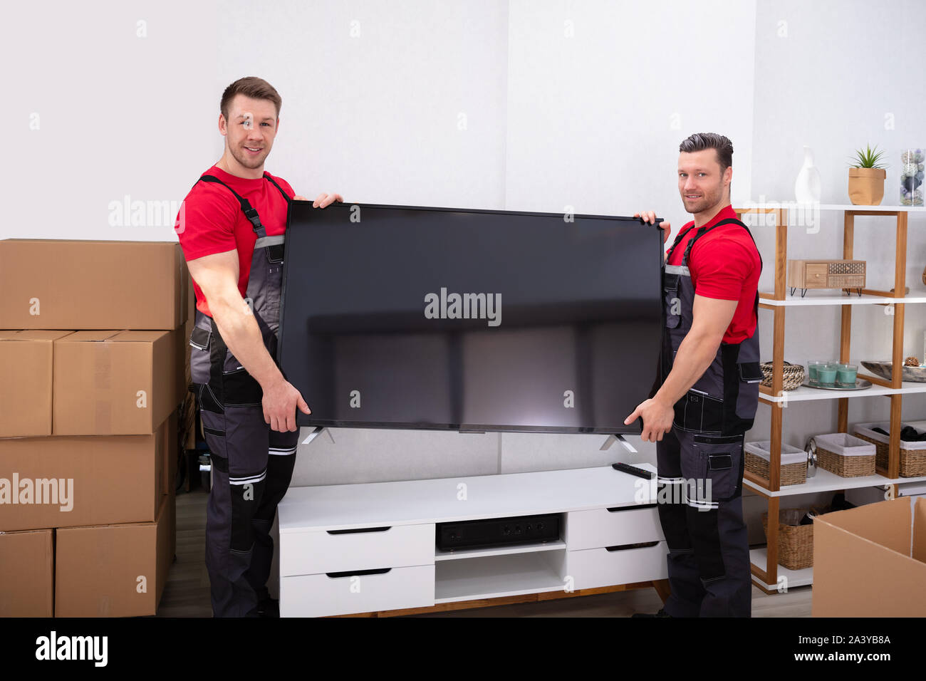 Two Male Professional Movers In Uniform Unloading LCD Television In The Living Room Stock Photo