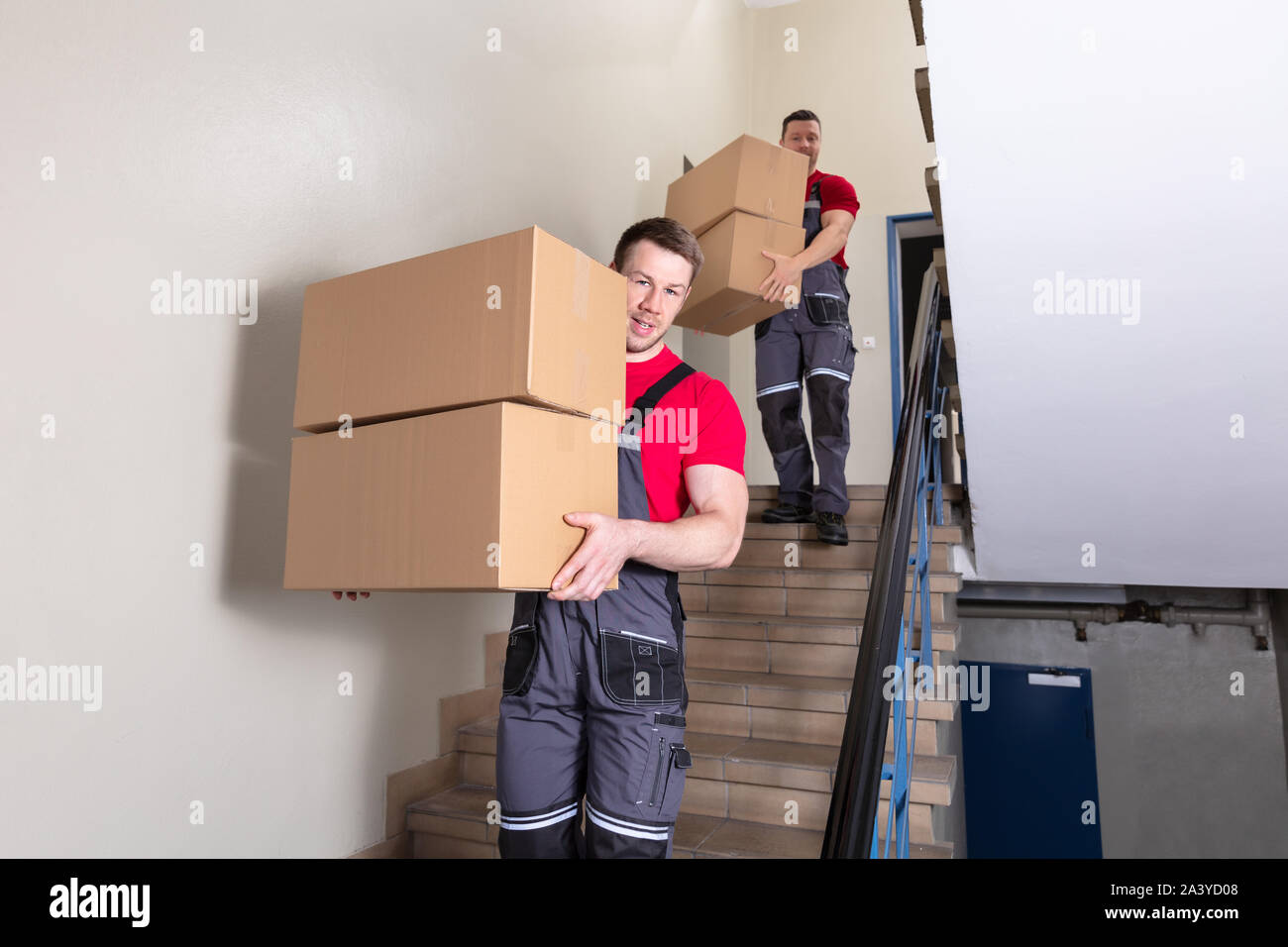 Portrait Of A Young Male Movers In Uniform Carrying Cardboard Boxes Walking Downward On Staircase Stock Photo