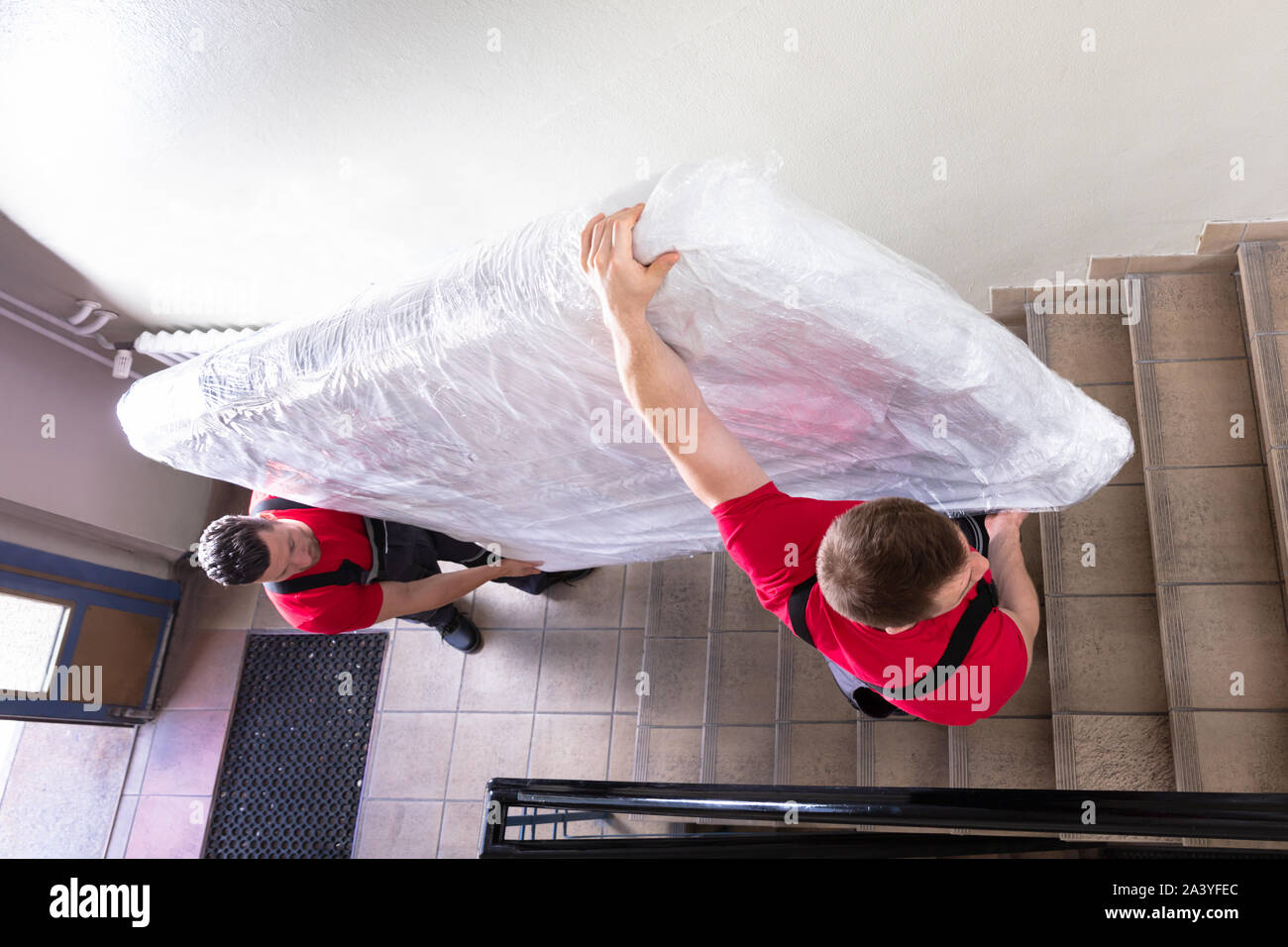 Two Young Male Movers In Uniform Carrying The Wrapped Mattress While