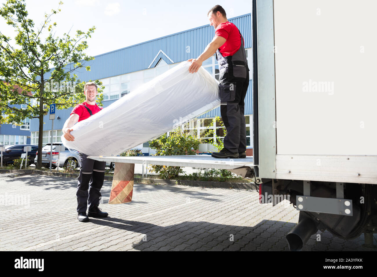 Young Male Movers Unloading The Wrapped Mattress From Moving Truck