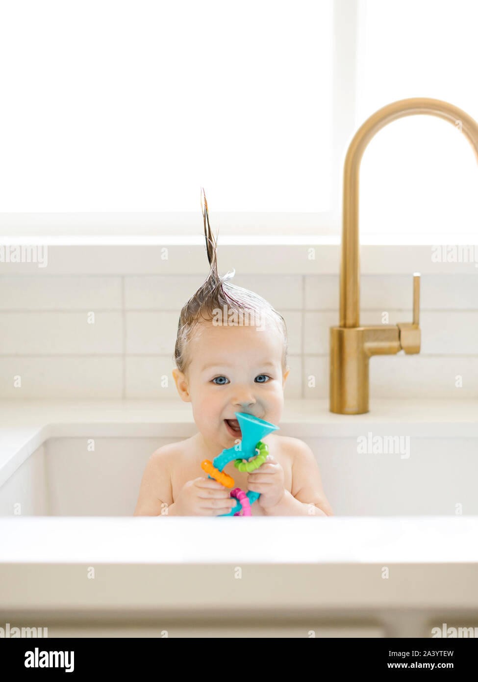 Baby girl with Mohican hairstyle holding toy while bathing in kitchen sink Stock Photo