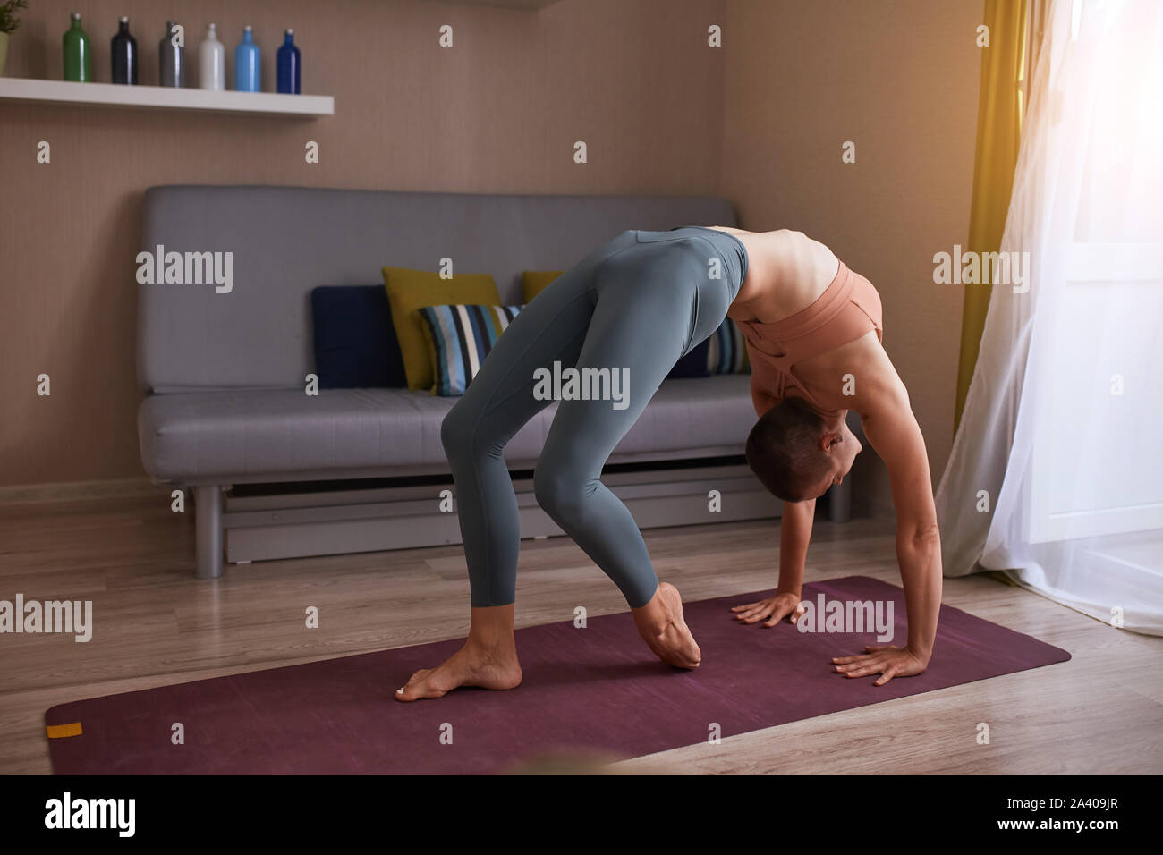 Young short-haired woman in grey leggins doing bridge exercise on purple mat, warming up muscles before practicing yoga at home Stock Photo