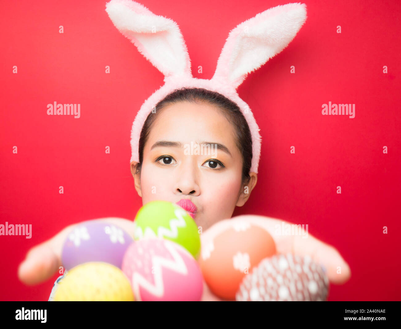 Woman wearing bunny ears headband and carrying Easter eggs during the Easter season. Attractive young woman and smiling on a bright red background. Stock Photo
