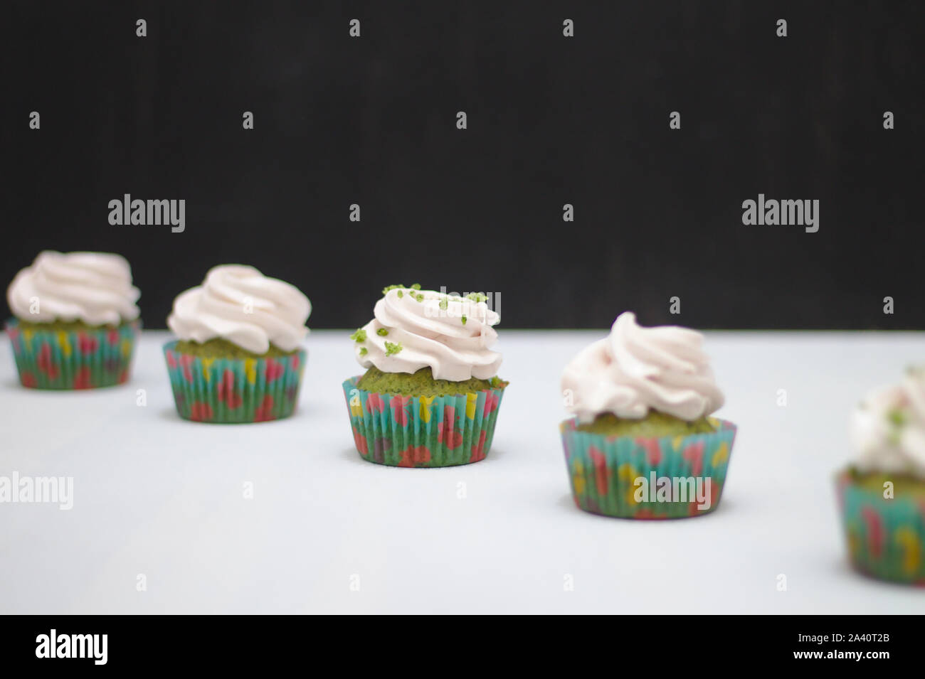 https://c8.alamy.com/comp/2A40T2B/green-cupcakes-with-cream-hat-in-multi-colored-papers-on-a-white-table-on-a-dark-background-focus-on-cake-depth-of-field-effect-2A40T2B.jpg