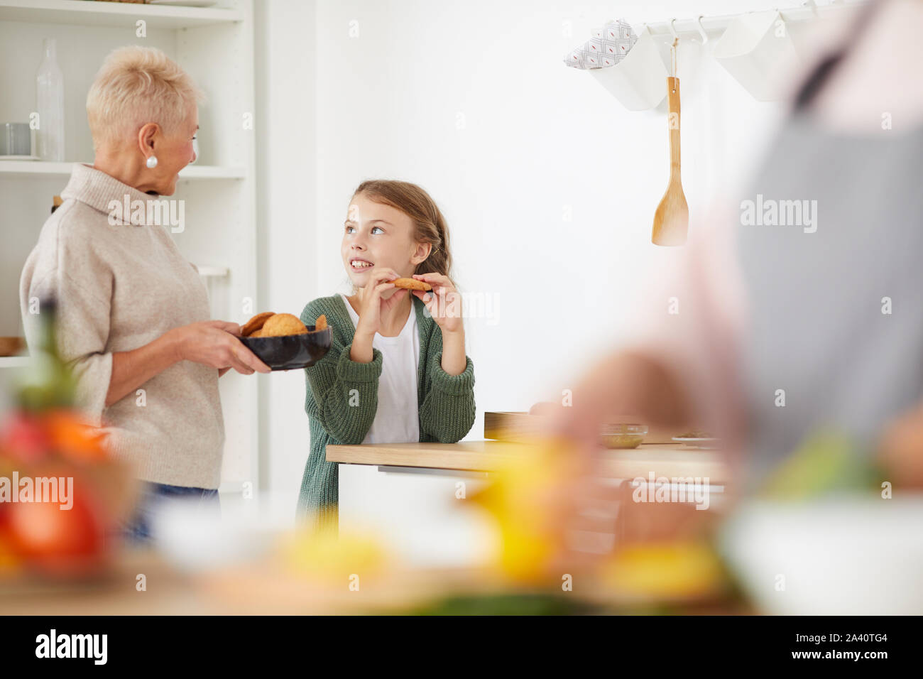 Little girl eating cookies and talking to her grandmother while they standing in domestic kitchen Stock Photo