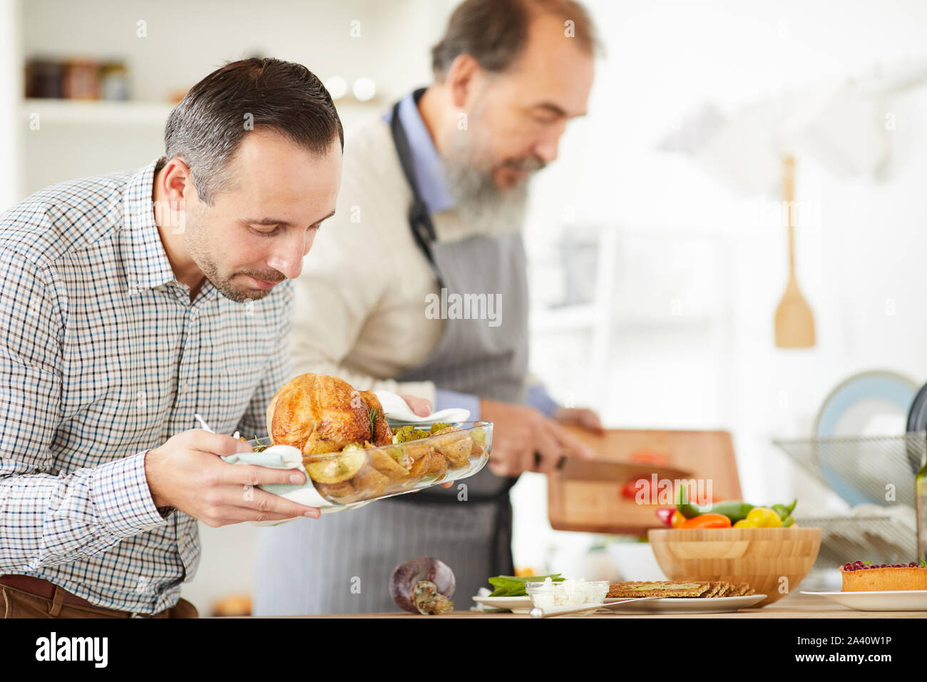 Young man looking at roast turkey in his hands with senior man cutting vegetable in the background at home Stock Photo