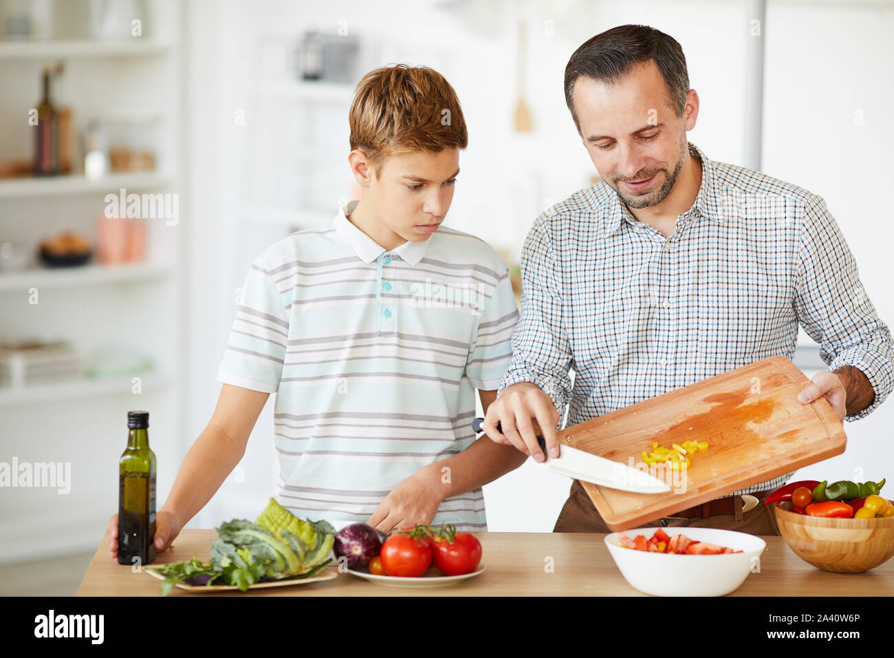 Young father cooking vegetable salad and teaching his son to cook in domestic kitchen Stock Photo