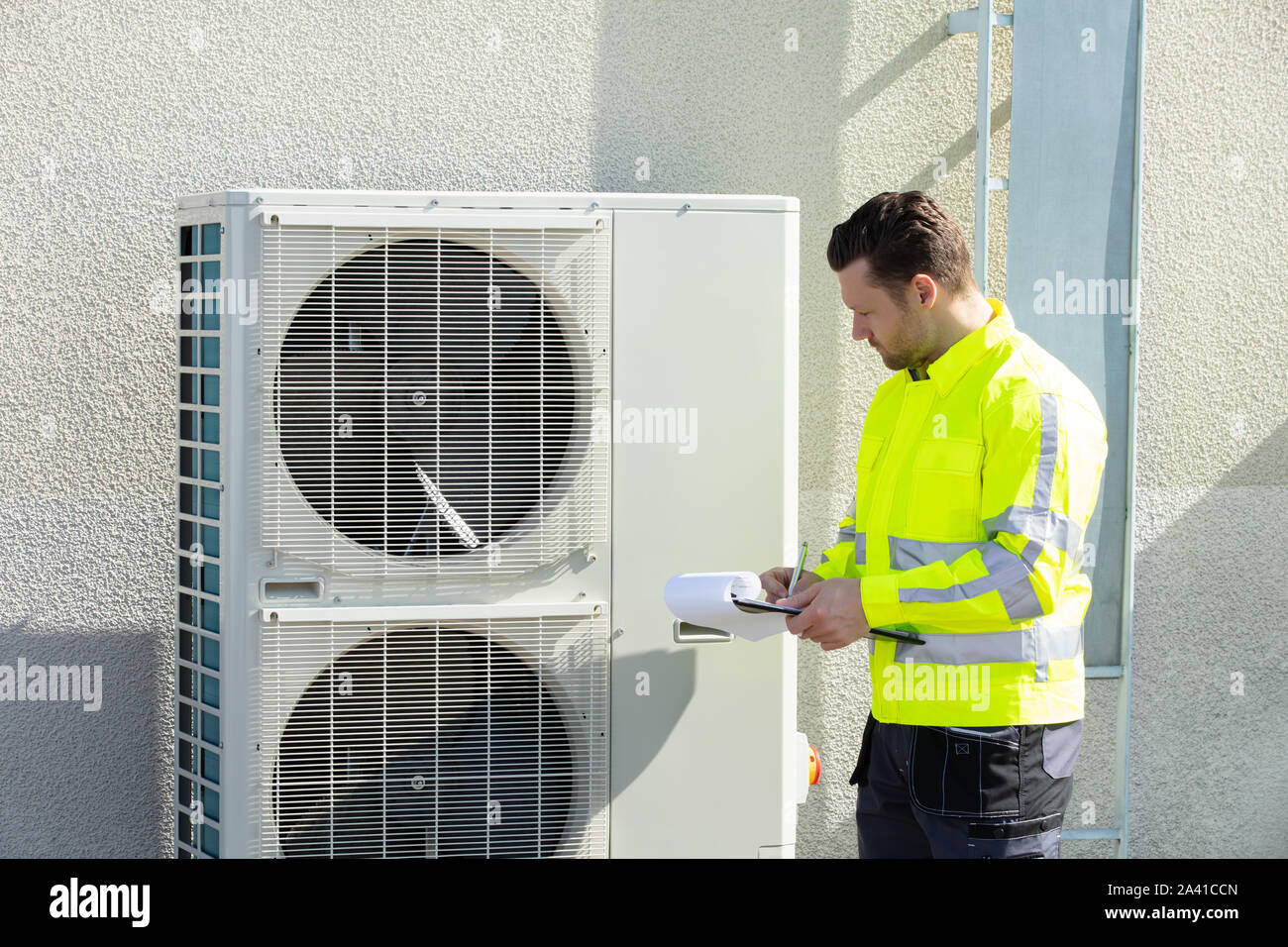 Young Male Technician Wearing Safety Jacket Looking At Air Conditioner Unit Making Notes Stock Photo