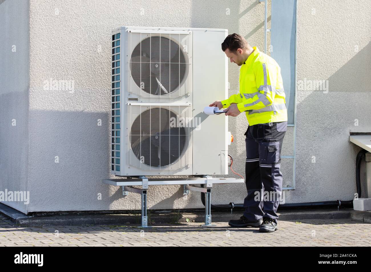 Young Male Technician Wearing Safety Jacket Looking At Air Conditioner Unit Making Notes Stock Photo