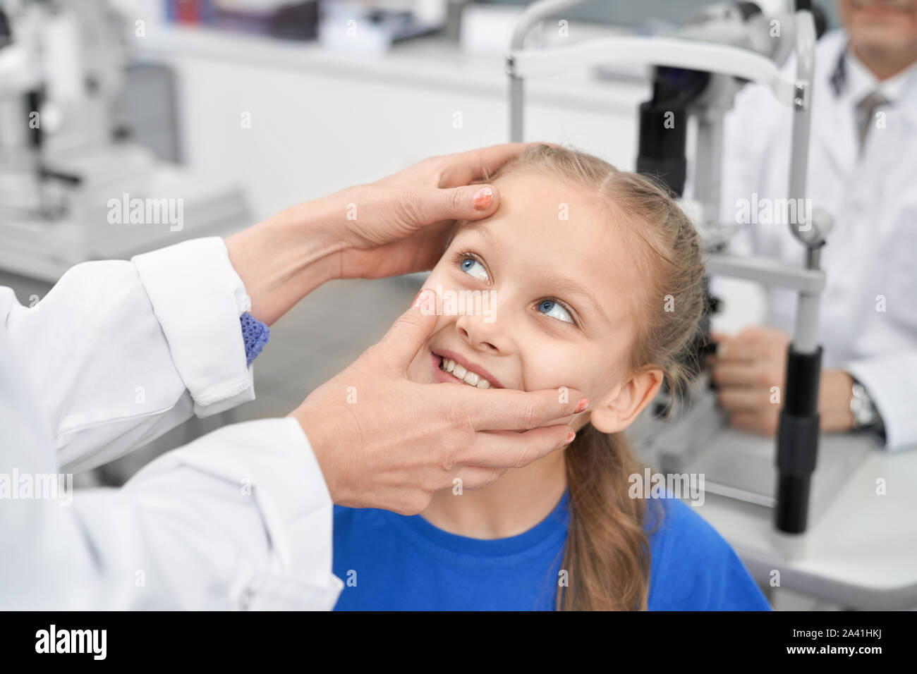 View from side of nice female oculist checking eye pupil of smiling girl in laboratory. Cheerful patient testing vision in clinic and choosing glasses or lenses. Concept of examination. Stock Photo