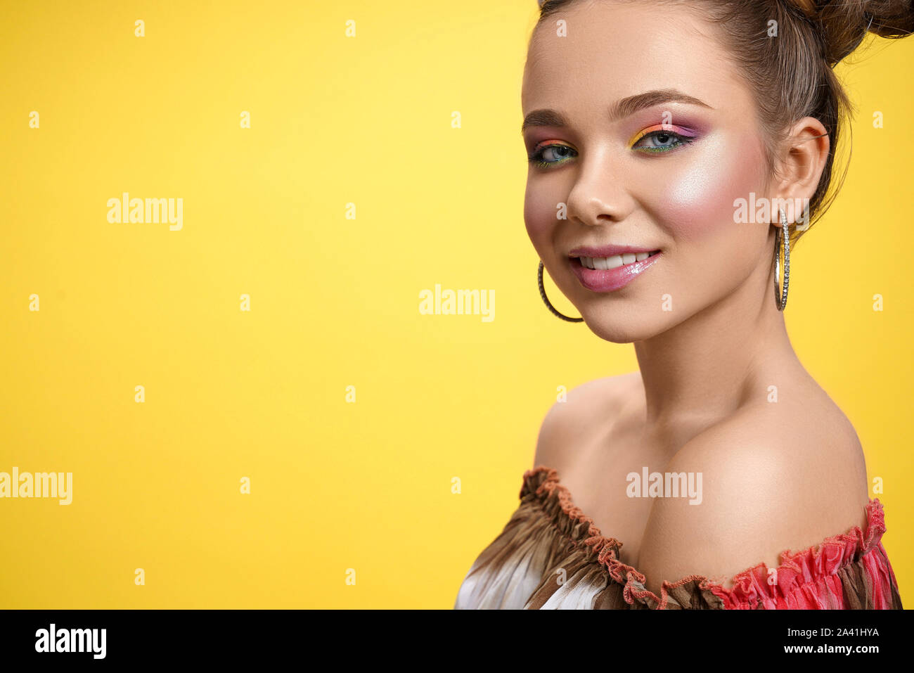 View from side of attractive female teenager with bright shadows looking at camera and smiling on yellow isolated background. Adorable girl with trendy hairstyle posing in studio. Concept of beauty. Stock Photo