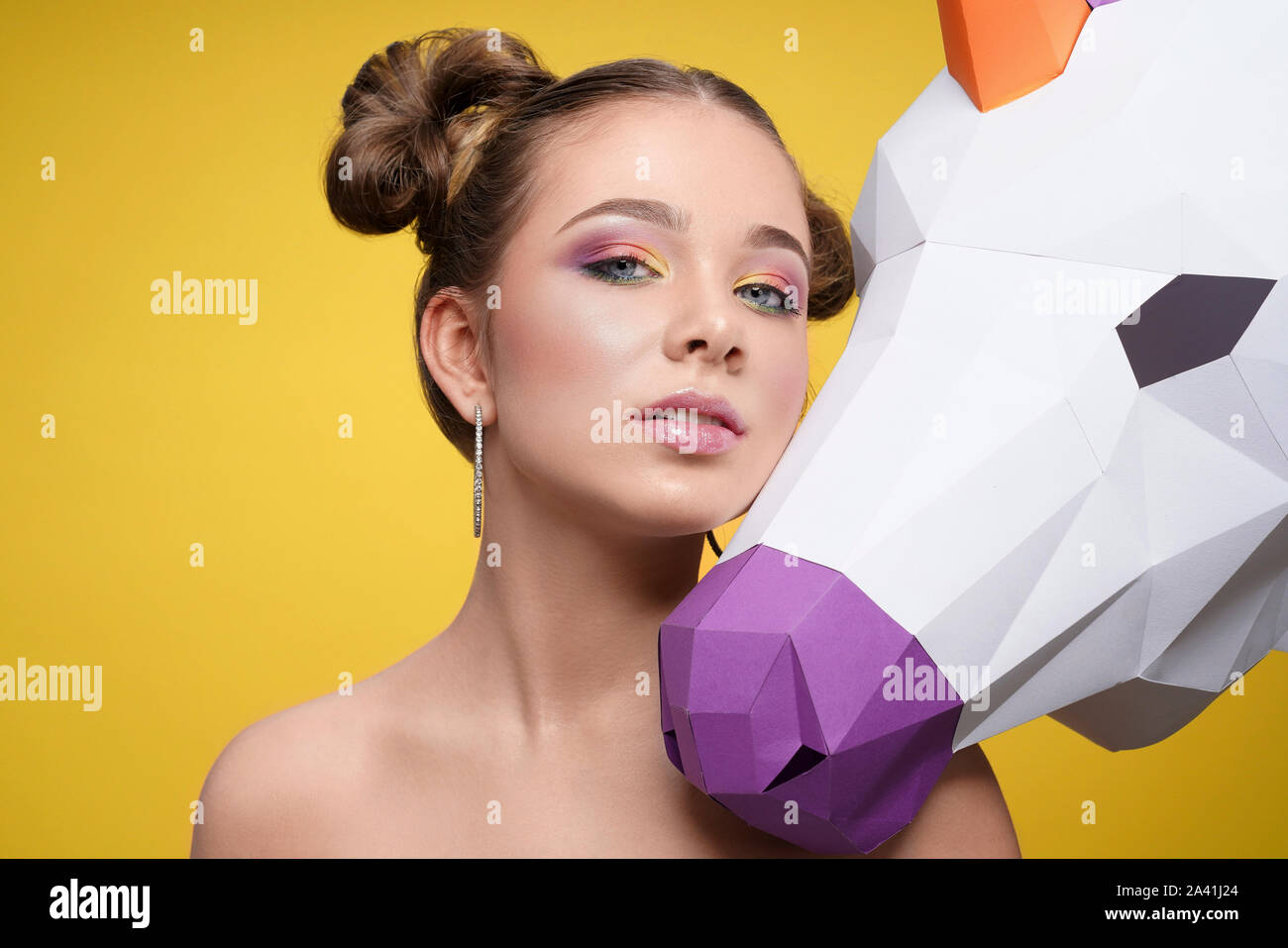 Front view of pretty young model with professional colorful makeup posing with 3d unicorn on yellow isolated background. Stylish girl with bright shadows looking at camera in studio. Stock Photo