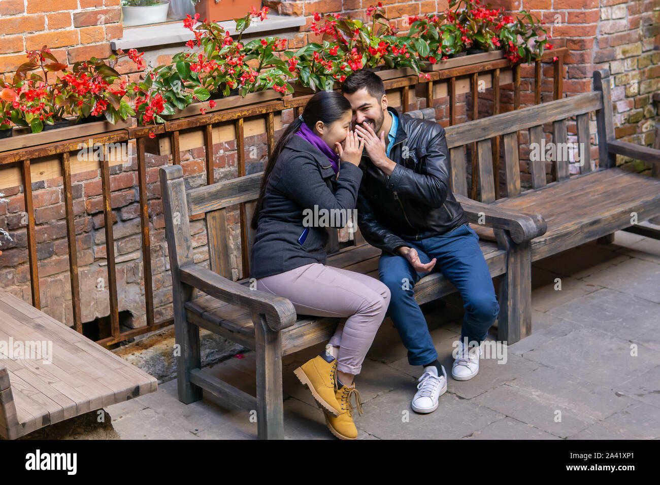 two young people a guy and a girl whisper about something and laugh sitting on a bench outdoors Stock Photo