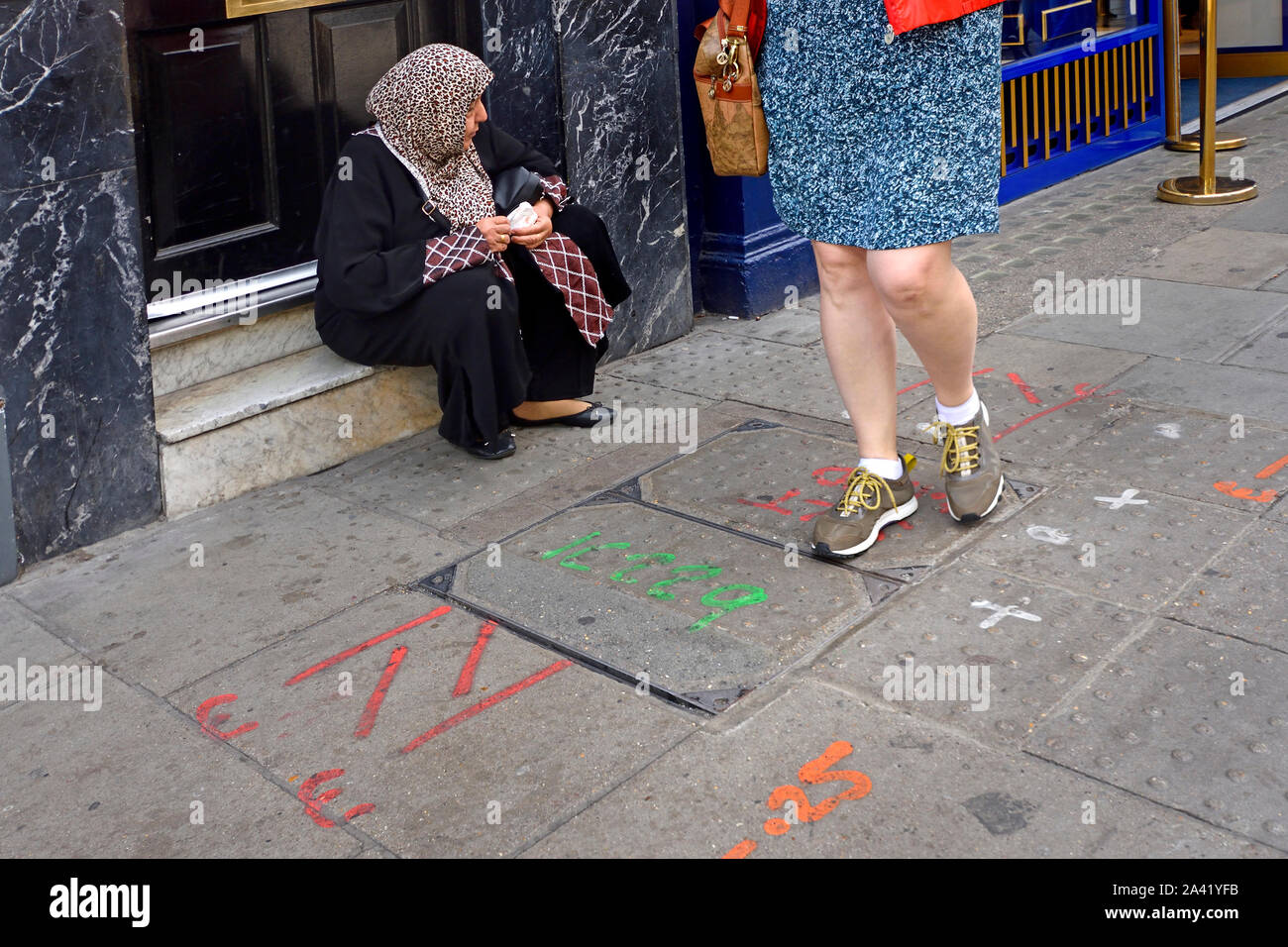 London, England, UK. Asian woman wearing a headscarf sitting in a doorway, a woman walking past Stock Photo