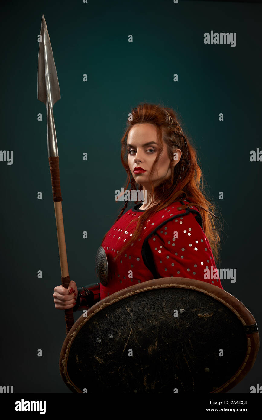 Side view of powerful female fighter in armor keeping spear and shield and posing on isolated background in studio. Young red haired woman looking at camera. Female hero fighting in combat. Stock Photo