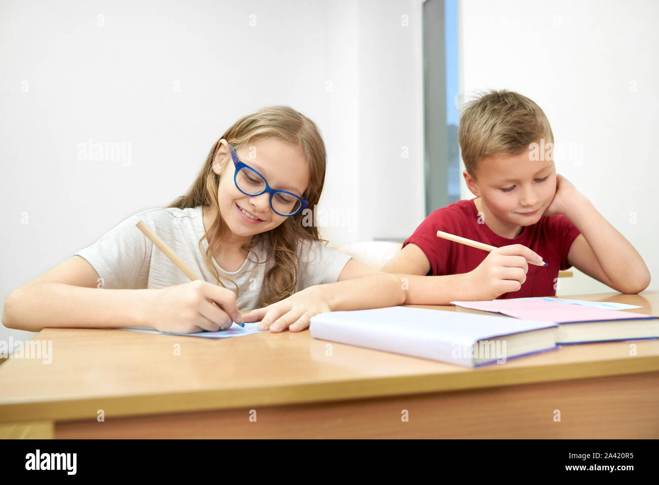 Smart students sitting in classroom and doing test at school. Pretty girl and boy keeping pen, looking at copybook and thinking in classroom. Little pupils in process of learning and studying. Stock Photo