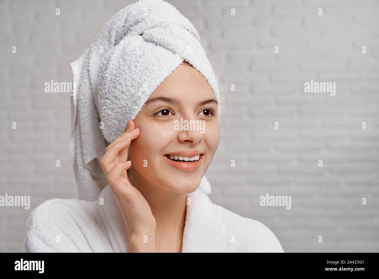 Cheerful, pretty woman with healthy, perfect skin of face posing, looking away. Beautiful girl using natural product for care of skin. Young woman in white bathrobe with white towel. Stock Photo
