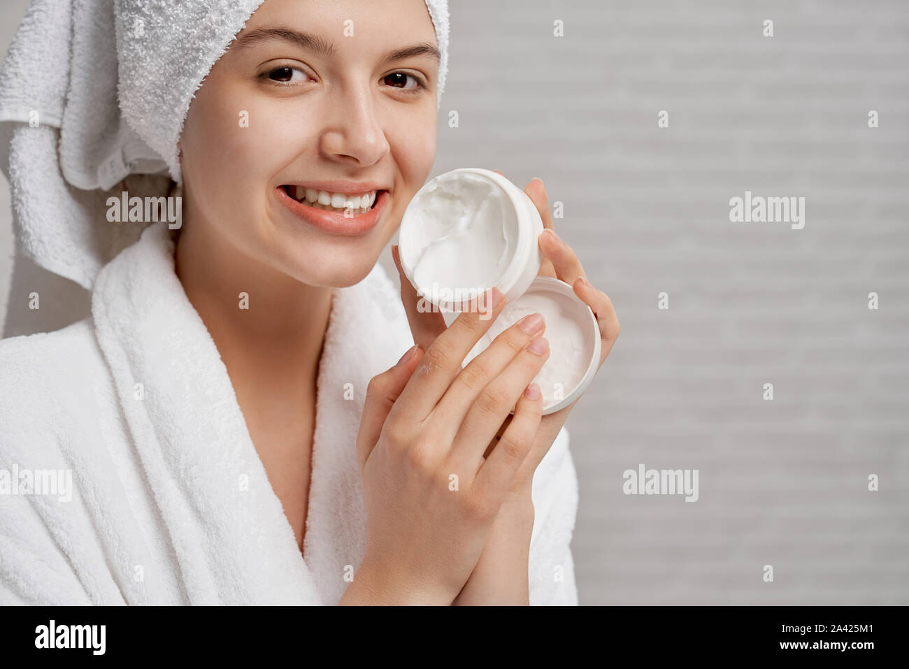 Pretty, young woman holding and showing white box with cosmetic cream. Beautiful model with perfect skin in white bathrobe and white towel on head smiling, looking at camera. Stock Photo