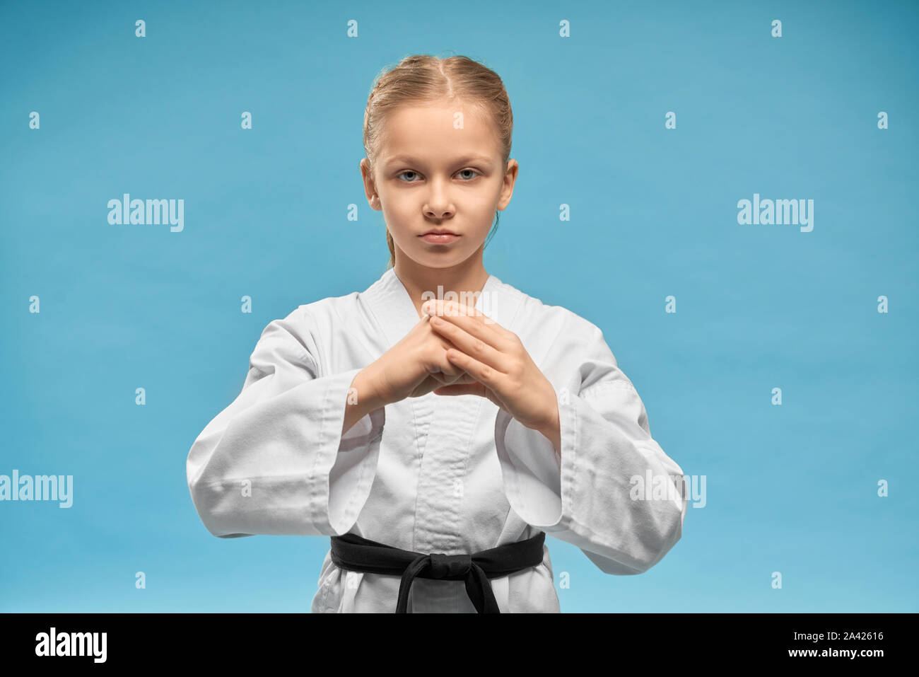 Young concentrated karate girl practicing hands position. Pretty junior in white kimono with black belt showing fist. Confident child posing on blue background, looking at camera. Stock Photo