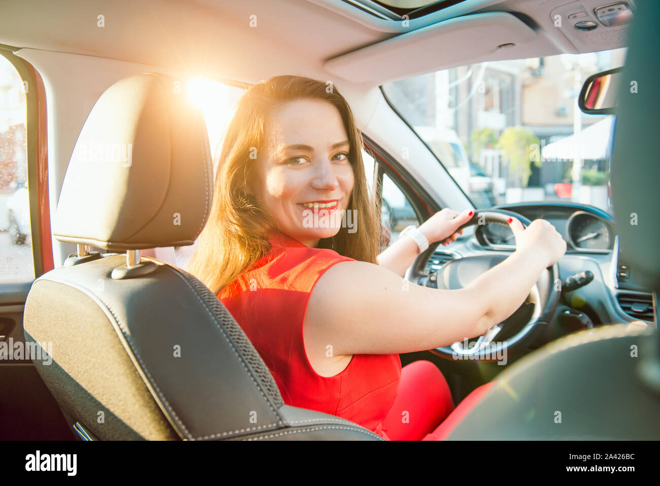 Back view Portrait of business lady, caucasian young woman driver looking at camera and smiling over her shoulder while driving a car. Sunset back lig Stock Photo