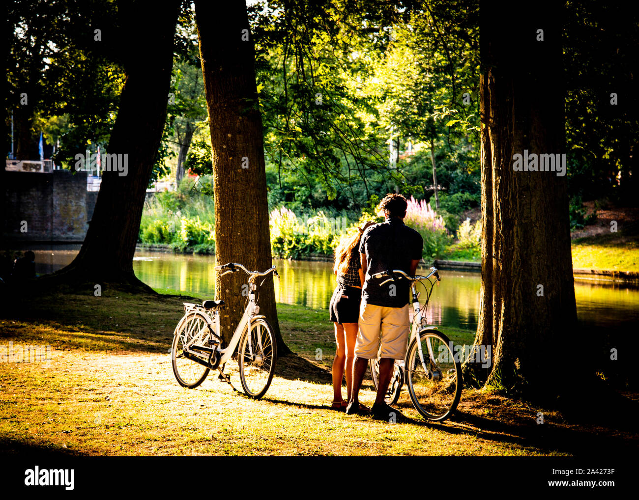 young couple standing together, backs to camera. taking a break,from bike ride as the setting sun paints atmospheric lighting across the canal banks. Stock Photo