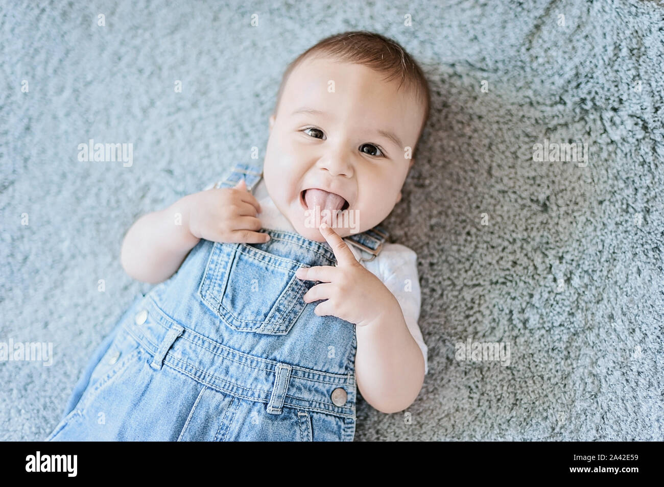 Funny little baby boy in denim lying on grey blanket. 6 month old child smiles with his tongue