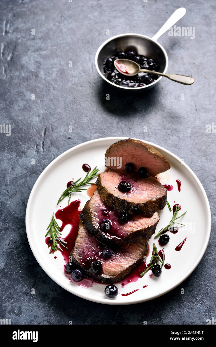 Tasty medium rare roast beef with berry sauce. Sliced grilled beef with blueberry sauce on white plate over blue stone background. Stock Photo