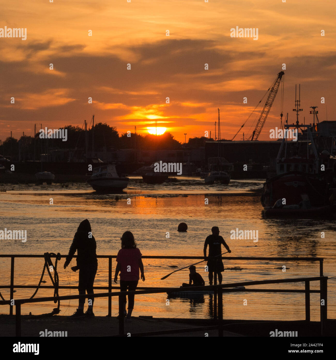 Young girls crabbing in the harbour at Leigh-On-Sea, silhouetted at sunset, as two paddle boarders set off. Stock Photo