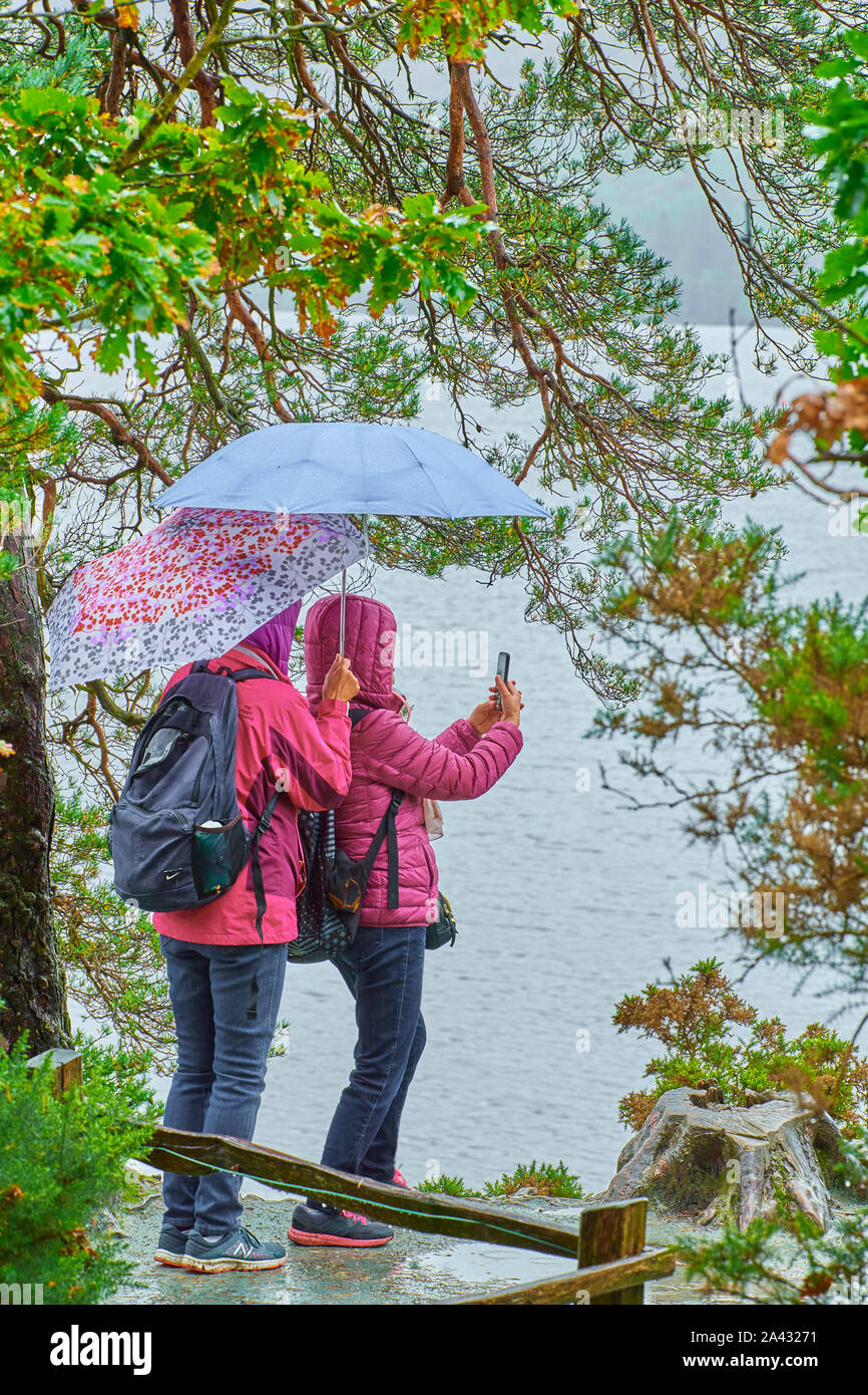 A couple of tourists with umbrellas take a selfie photo on the shoreline during a cloudy, rainy day at Lake Derwentwater, Lake District national park, Stock Photo