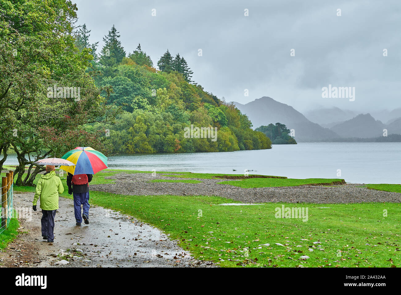 A man and a woman with their umbrellas up walk on a path by the shore on a cloudy, rainy day at Lake Derwentwater, Lake District national park, Cumbri Stock Photo