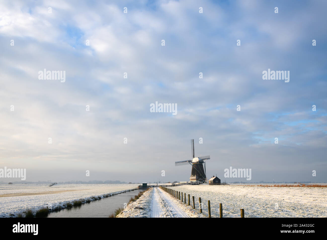 View on the dutch countryside in winter with a path with a fence and a ditch towards a windmill, covered in snow under nice clouds in the Netherlands. Stock Photo