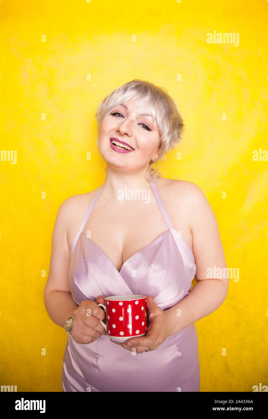 pretty curvy young blonde woman with short hair enjoys drinking tea from  cute red polka dot ceramic cup on yellow studio background Stock Photo -  Alamy