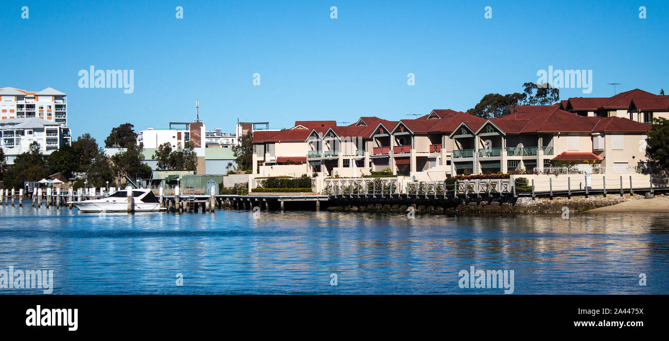 Large waterside houses, apartment condominiums in suburban community on riverfront with boats moored at wharf, blue sky in background Stock Photo