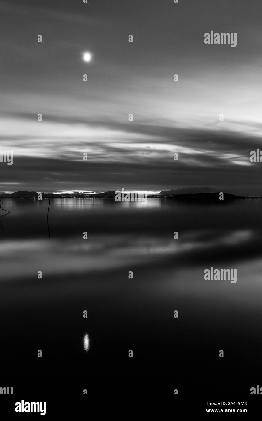 Moon above a lake with its reflection on the Trasimeno lake surface at dusk Stock Photo