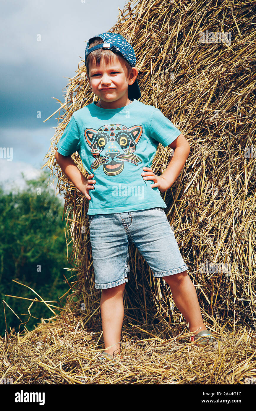 A little boy in a blue baseball cap and short denim shorts plays on straw bales on a hot summer day Stock Photo